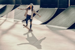 Süßes gemischtrassiges Teenagermädchen mit lockigem Haar und Mütze, das Skateboard im Skatepark fährt.