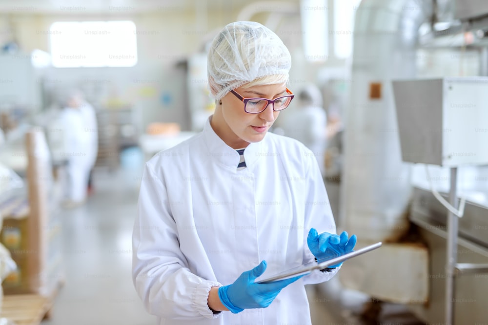 Supervisor in sterile uniform and with eyeglasses using tablet for controlling workflow in food factory.