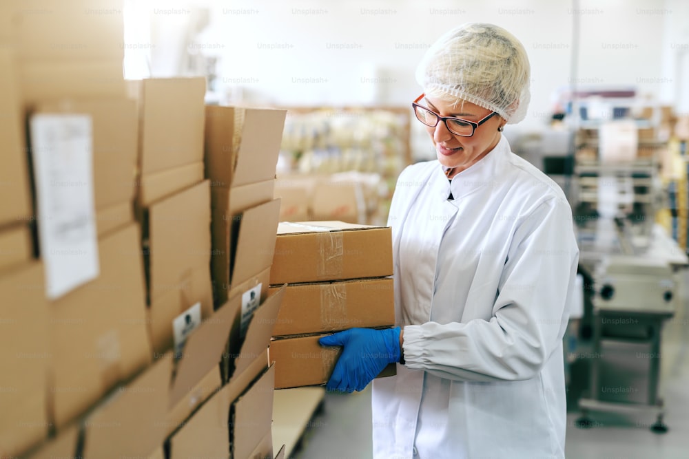 Smiling female blonde employee in sterile uniform and with eyeglasses putting boxes on stack. Food factory interior.