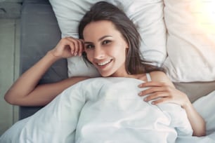 Top view of beautiful young woman is lying on bed, smiling and looking at the camera