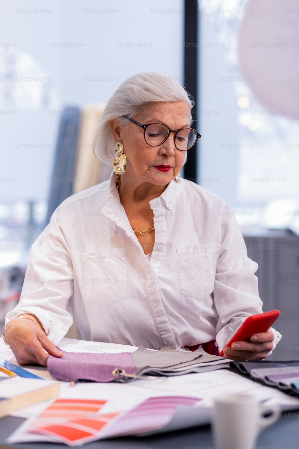 몇 년 만에 비즈니스 마담. 노년의 위엄 있는 비즈니스 마담과 흰색 블라우스, 거대한 황금 귀걸이, 사무실 테이블에 앉아 전화로 교환을 확인하는 생생한 컬러 립스틱