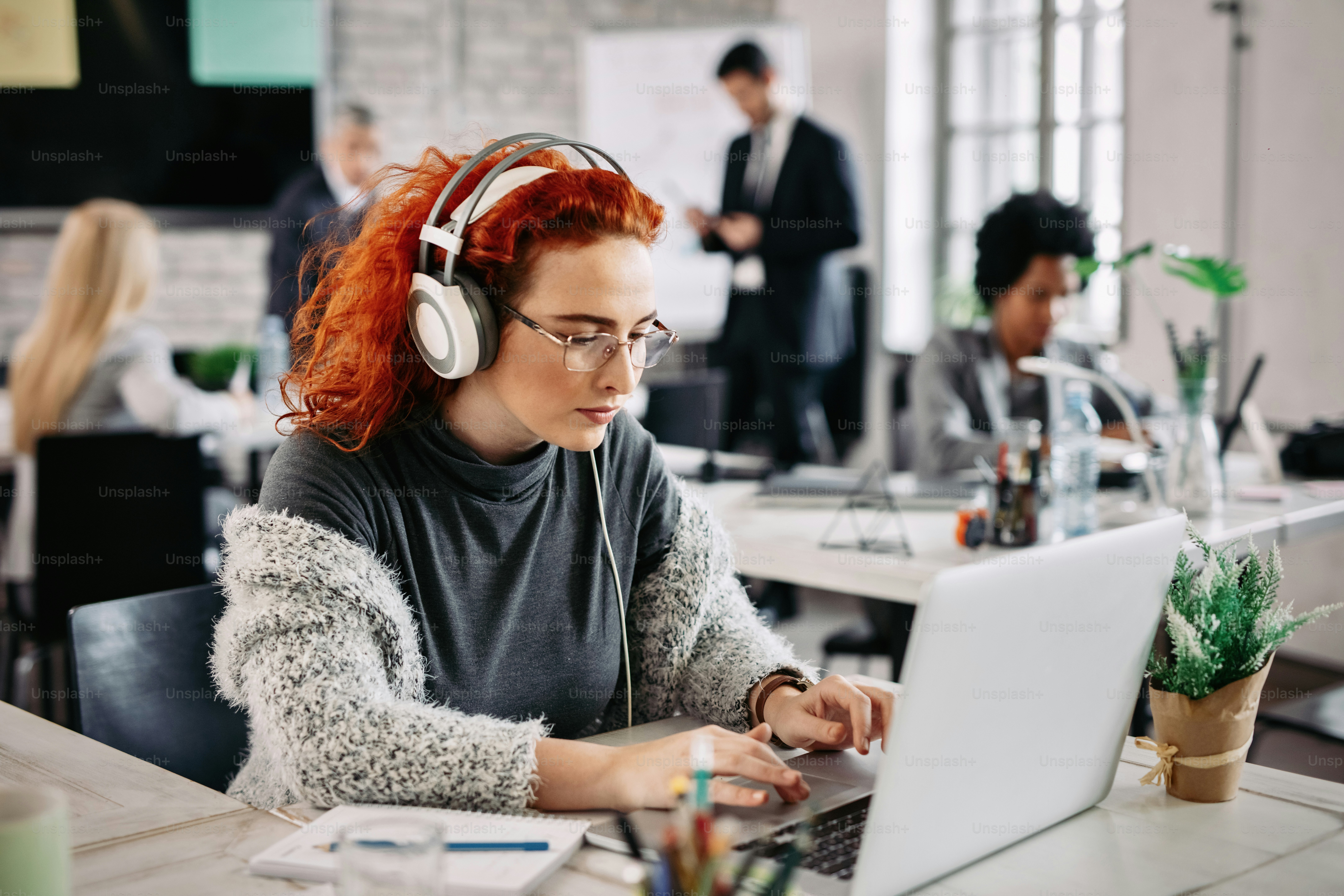 Young businesswoman surfing the net on laptop while listening music on headphones and working in the office. There are people in the background.