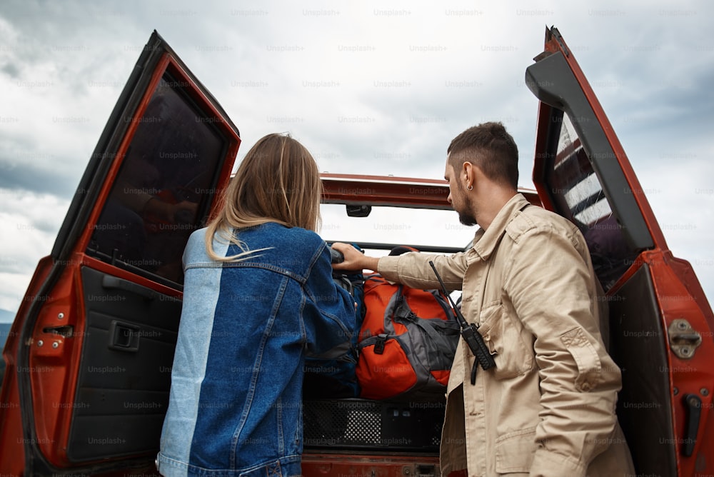 Rear view of a young couple of travelers having an active weekend while standing near trunk of their off-road vehicle