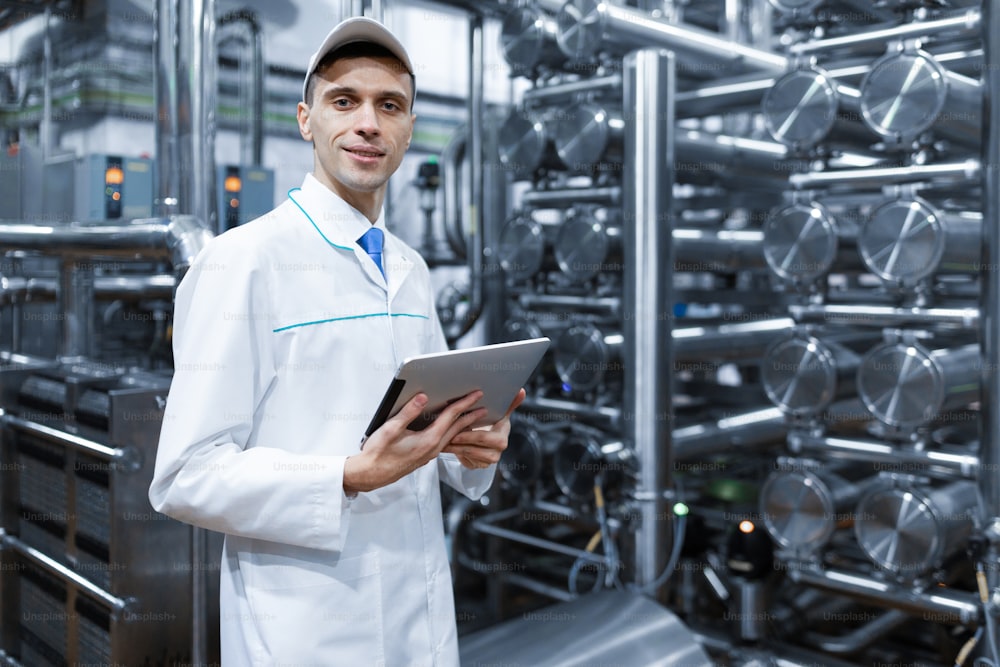 Technologist in a white coat makes the necessary entries in the tablet is at the factory. A man with a digital tablet in uniform in the production shop. interior of production department on background