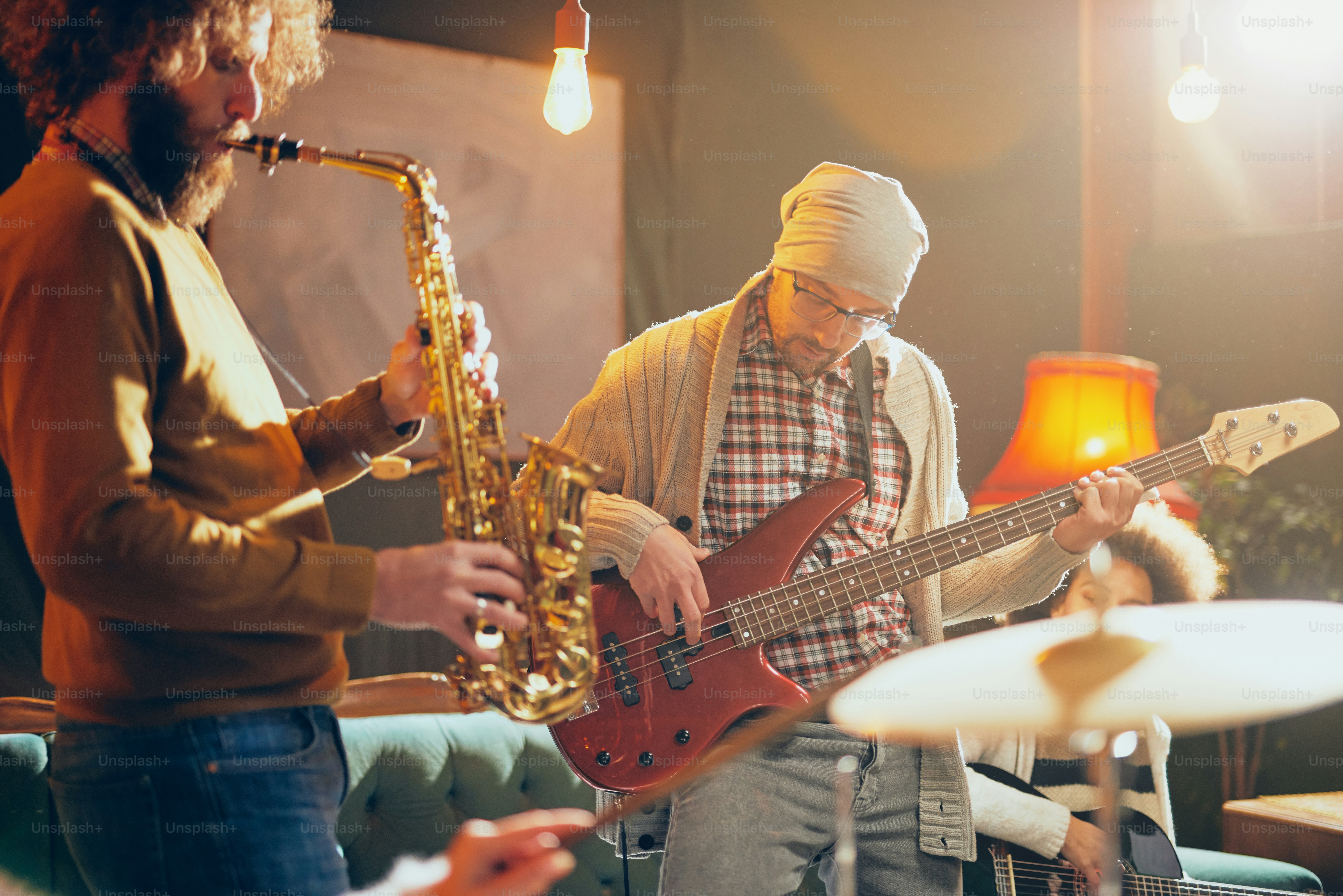 Young Caucasian man with hat on head playing bass guitar while sitting in home studio. In background saxophonist playing his instrument.