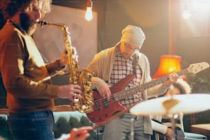 Young Caucasian man with hat on head playing bass guitar while sitting in home studio. In background saxophonist playing his instrument.