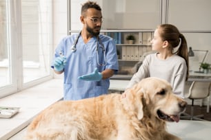 Young veterinary professional in gloves and uniform preparing syringe with medicine and consulting little owner of labrador dog