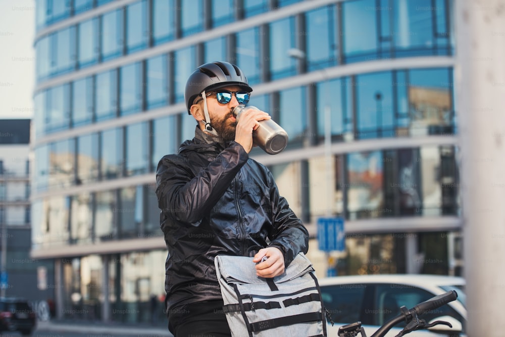Un corriere maschio con occhiali da sole che beve acqua durante la consegna di pacchi in città.