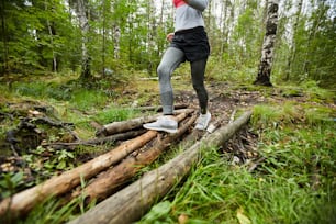 Young sportswoman in activewear moving down bunch of logs on forest path while training there in the morning