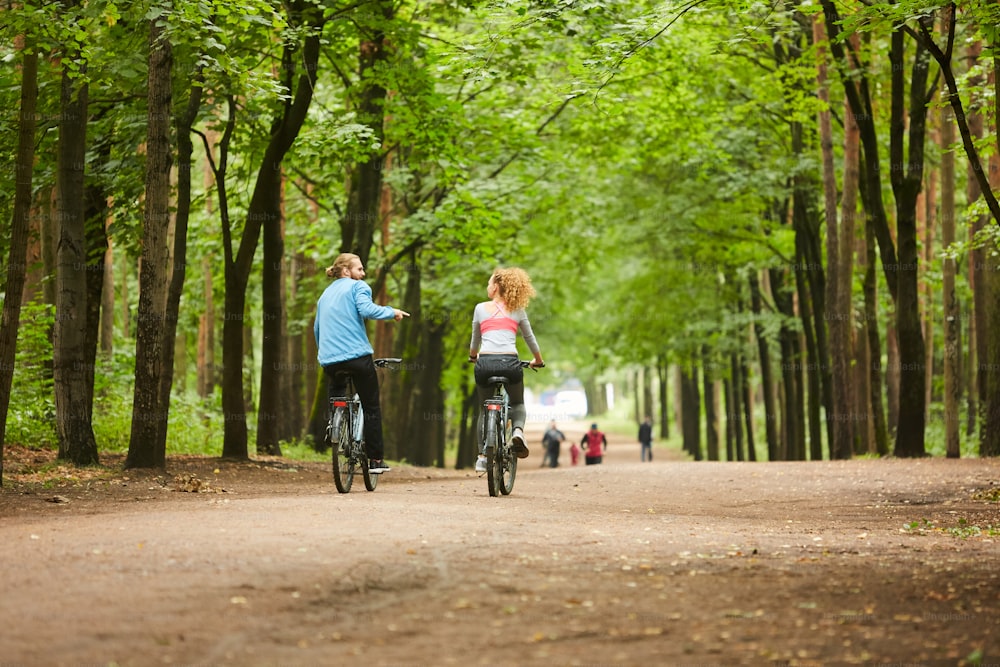 Rückansicht eines jungen Paares in Aktivkleidung, das sich auf Fahrrädern bewegt und am Sommertag im Park mit Menschen plaudert, die vor ihnen laufen