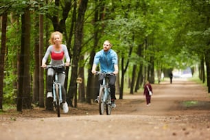 Zwei junge aktive Radfahrer fahren an einem Sommertag auf Fahrrädern auf einer breiten Straße zwischen Bäumen im Park