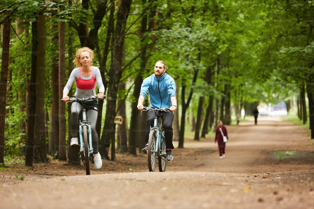 Dos jóvenes ciclistas activos montando en bicicletas a lo largo de un camino ancho entre árboles en el parque en un día de verano
