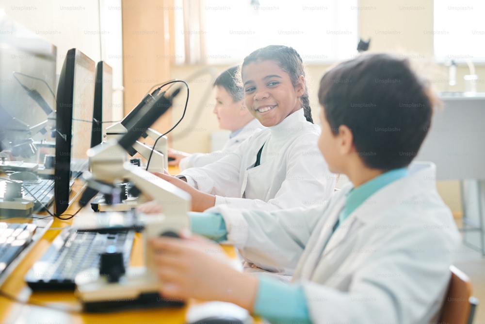 Happy little schoolgirl looking at her classmate at lesson of chemistry while working with microscope