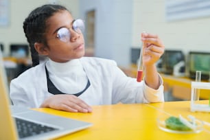 Serious clever schoolgirl in eyeglasses and whitecoat holding flask with chemical substance inside and looking at it