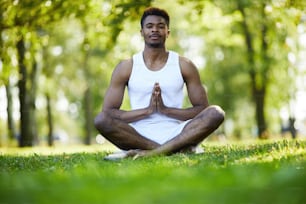 Contento joven negro guapo con bigote sentado con las piernas cruzadas y tomados de la mano en Namaste mientras practica yoga en el parque de verano