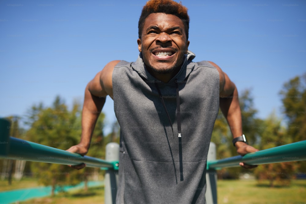 Aggressive emotional handsome young black man with clenched teeth doing dips on metal parallel bars outdoors
