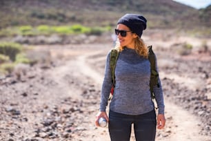 Cheerful cute curly woman with hat and trekking hiking clothes smile during a walk at the mountain - alternative outdoor leisure activity for caucasian people - standing beautiful woman
