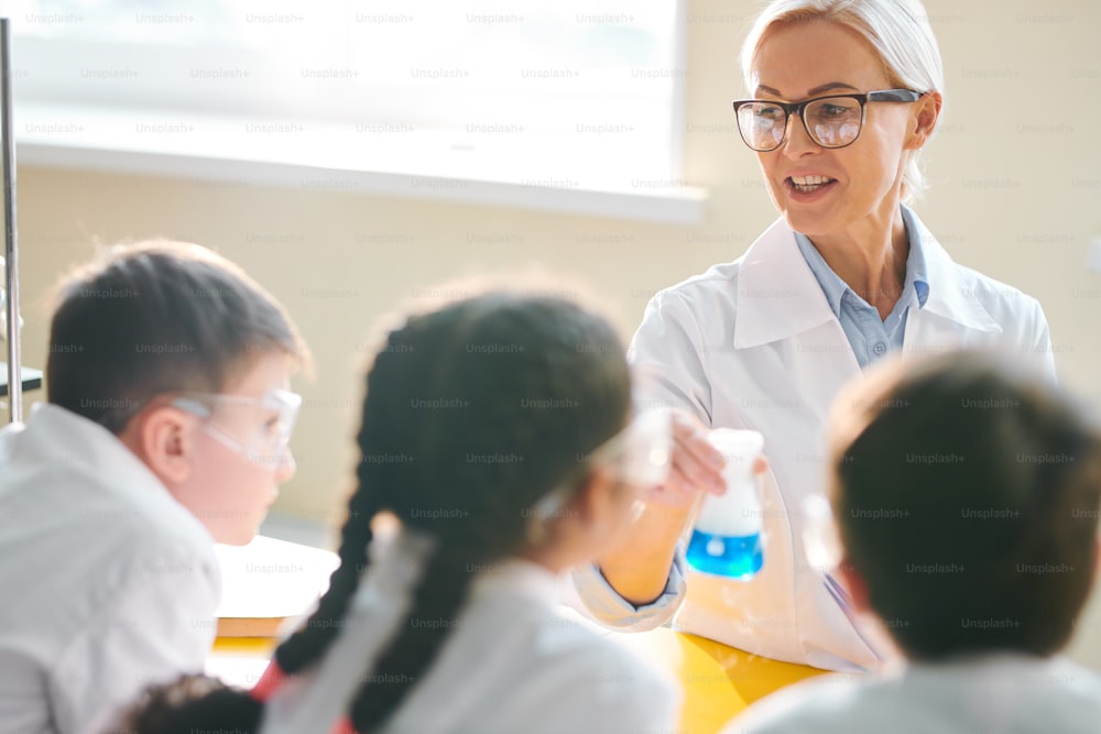 Blonde mature teacher in eyeglasses holding beaker with blue fuming fluid while explaining students what is happening