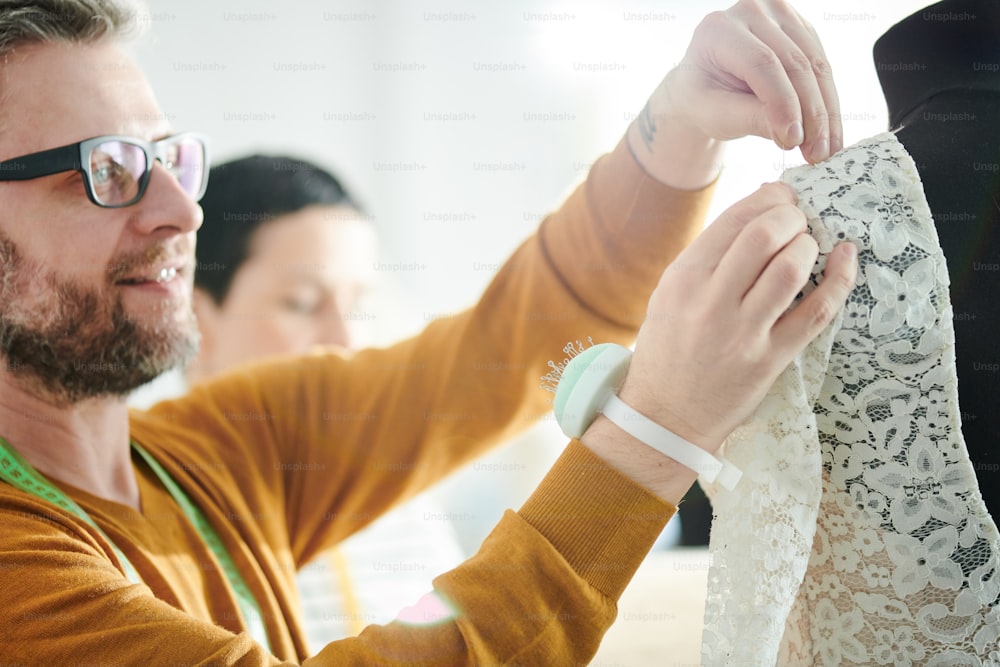 Reife Schneiderin in Brille und Pullover Pinning Ärmel aus Spitze Brautkleid auf Schnuller in der Werkstatt