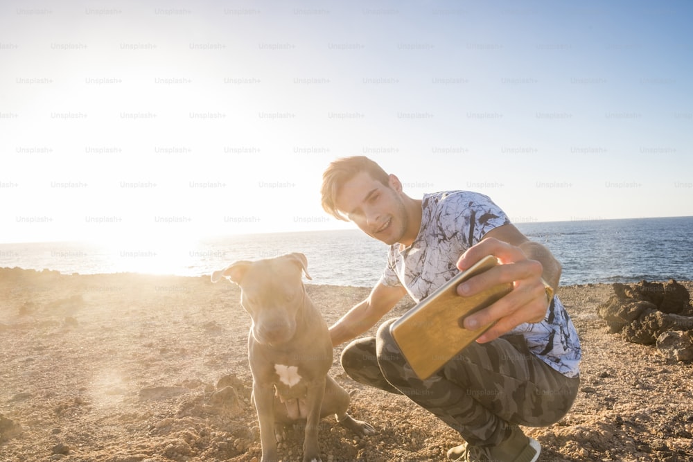 Happy couple of alternative millennial friends man and dog taking selfie picture with modern phone during a sunset in outdoor leisure activity together neat the ocean - sunny backlight and pet therapy lifestyle