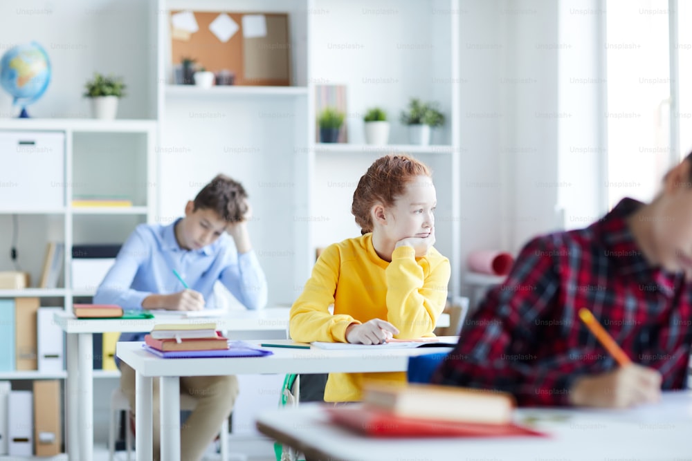 One of schoolkids sitting by desk among classmates and looking through classroom window