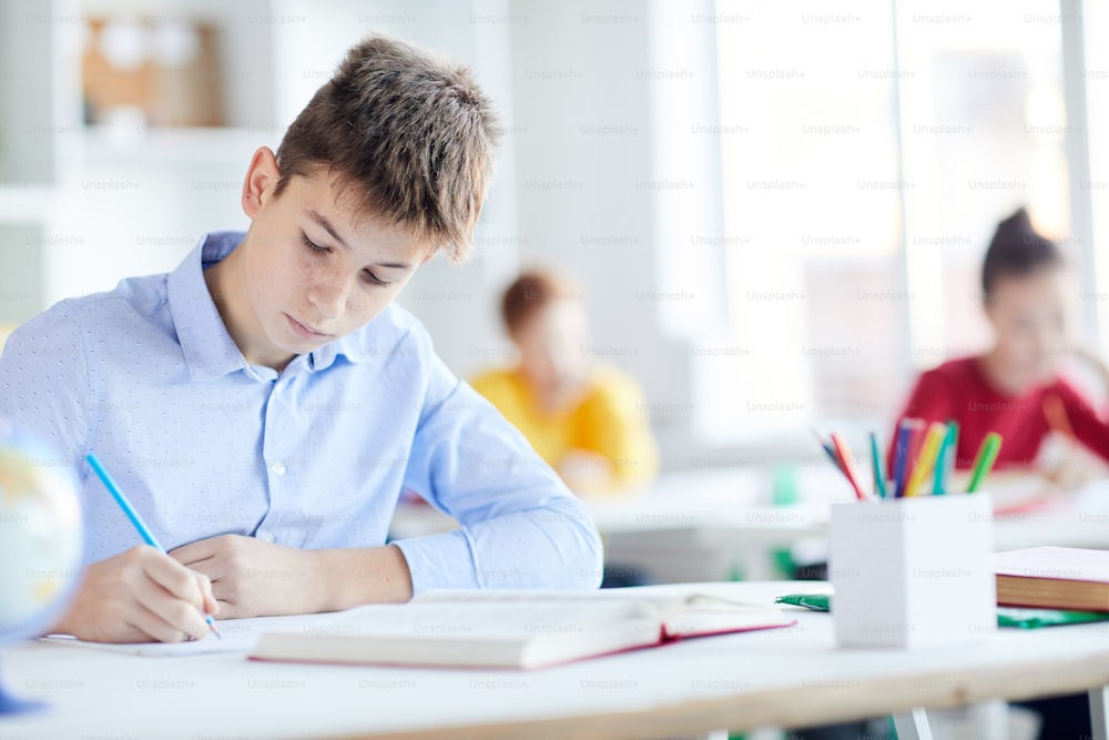 Serious schoolboy with pencil making notes on paper with open book in front and classmates behind