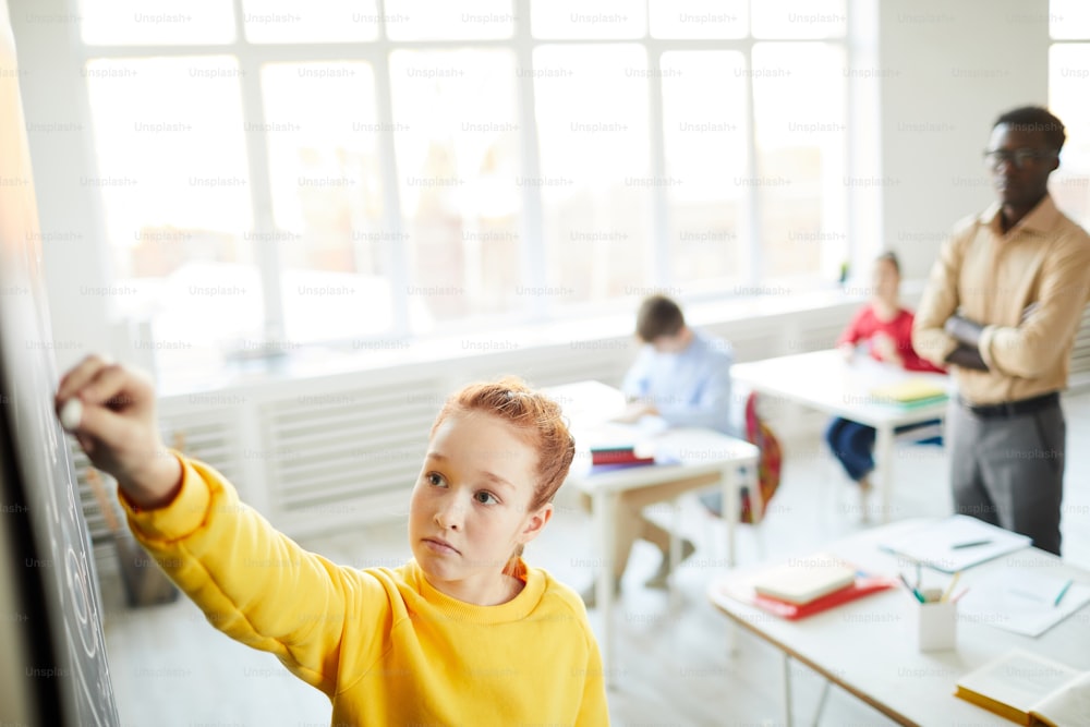 캐주얼 복장을 입은 영리한 여학생이 칠판 옆에 서서 수업 시간에 교실 앞에서 분필로 글을 씁니다.