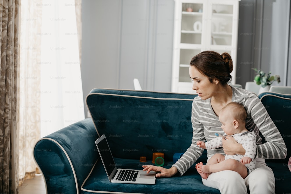I work on maternity leave. Beautiful lady keeping her child in hands and typing on computer at home. She is sitting on sofa in living-room indoors