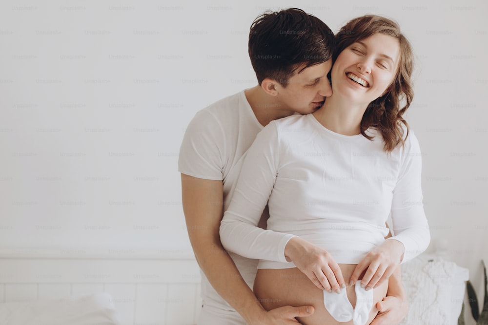 Happy young husband kissing his pregnant smiling wife and holding white little socks on belly bump. Stylish pregnant family, mom and dad in white, relaxing at home and hugging belly.