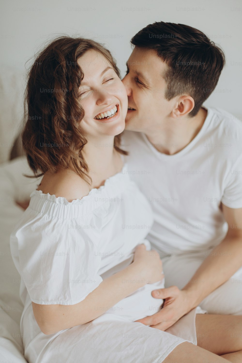 Glücklicher junger Ehemann, der seine lächelnde schwangere Frau küsst und Bauchstoß auf weißem Bett umarmt. Stilvolles schwangeres Paar in Weiß entspannt sich und hält den Bauch zu Hause. Fruchtbarkeitskonzept. Wahres Glück