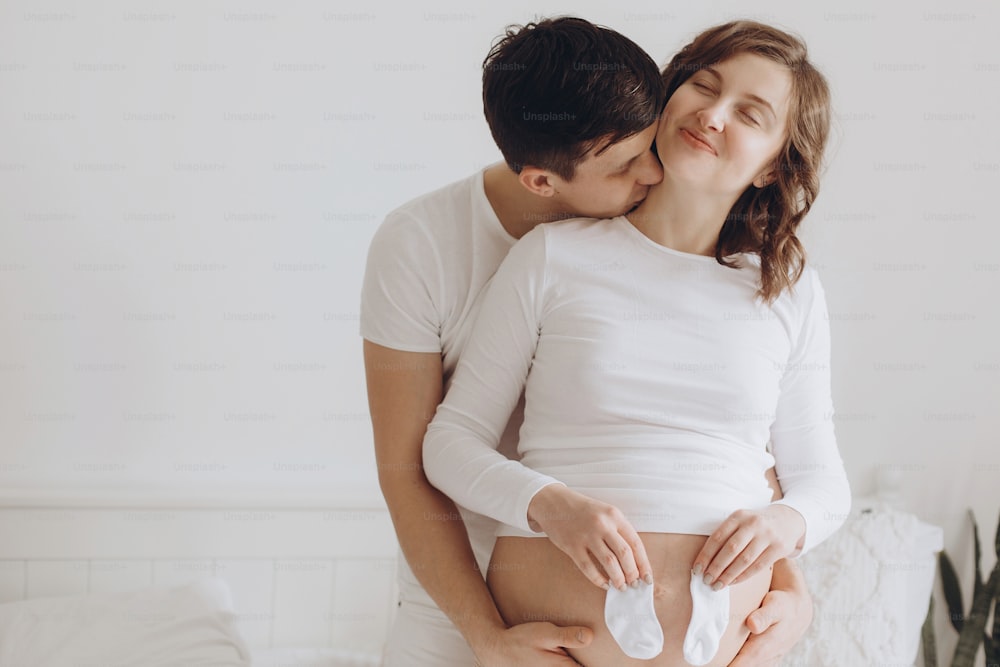Happy young pregnant couple holding white little socks on belly bump on white bed. Stylish pregnant family, mom and dad in white, relaxing at home and hugging belly. Fertility concept.
