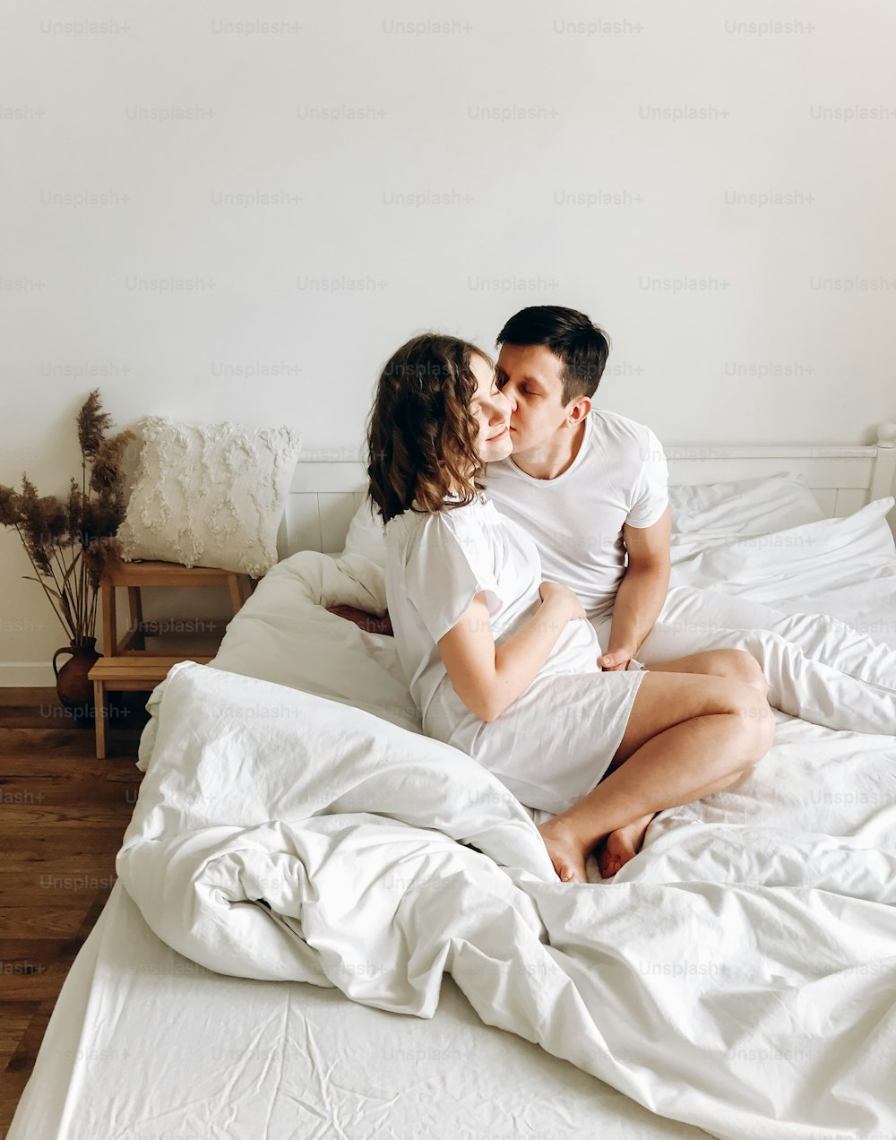 Happy young husband kissing his smiling pregnant wife and hugging belly bump on white bed. Stylish pregnant couple in white relaxing and holding belly at home. Fertility concept. True happiness