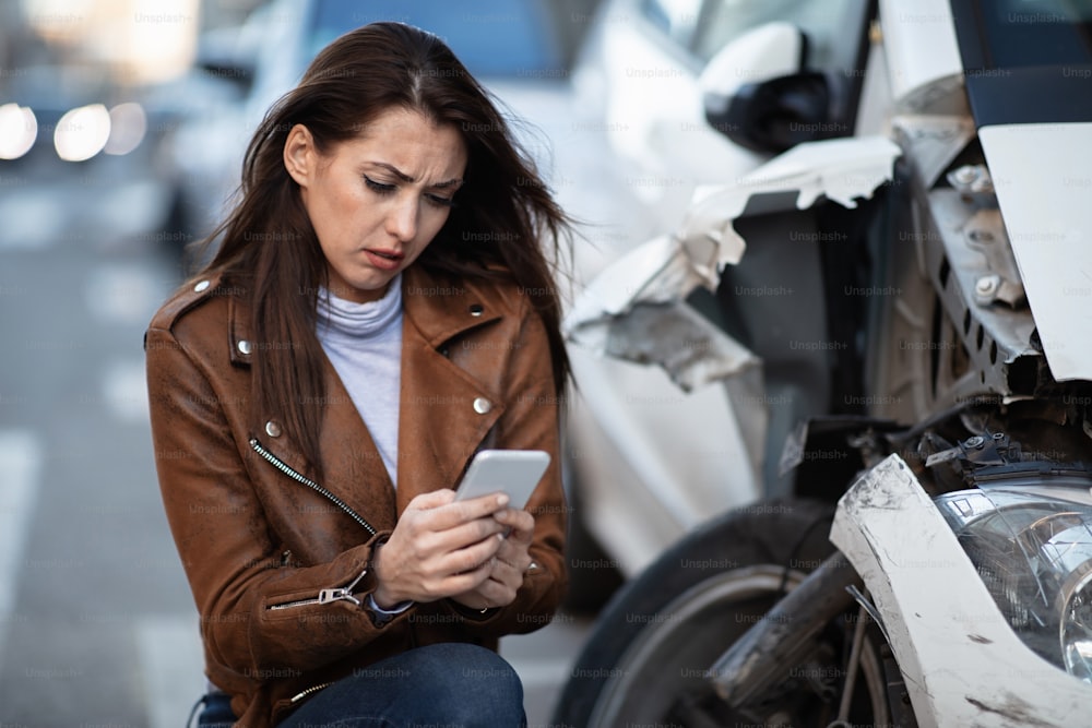 Mujer desesperada escribiendo en el teléfono móvil mientras está al lado de su coche destrozado después del accidente.