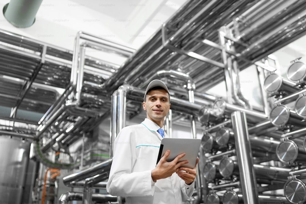 Technologist in a white coat makes the necessary entries in the tablet is at the factory. A man with a digital tablet in uniform in the production shop. interior of production department on background