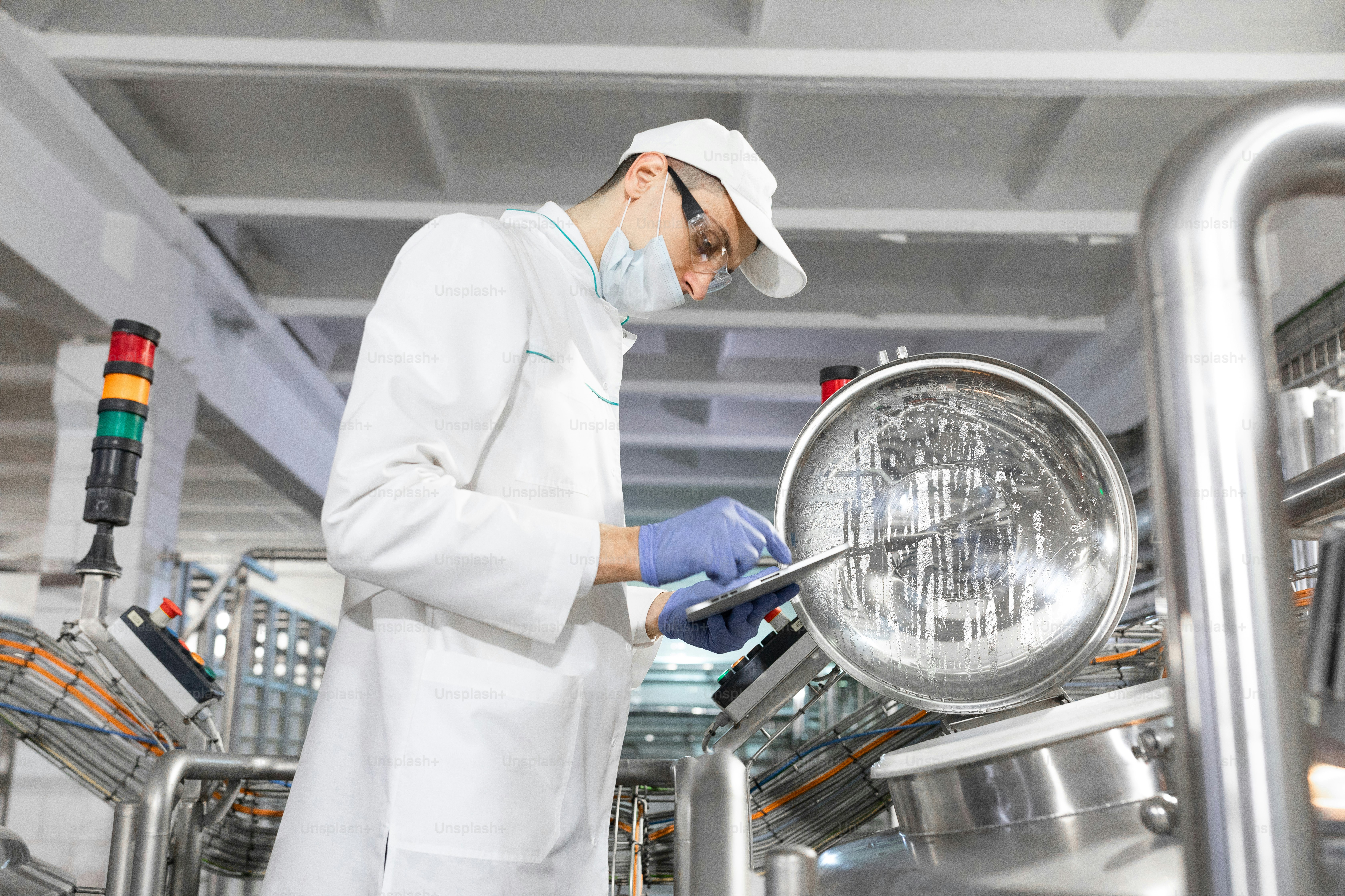 Technologist in a cap, mask and white coat is in the production shop and holding a digital tablet. inspector removes the indicators at the dairy plant. Engineer keeps statistics on production