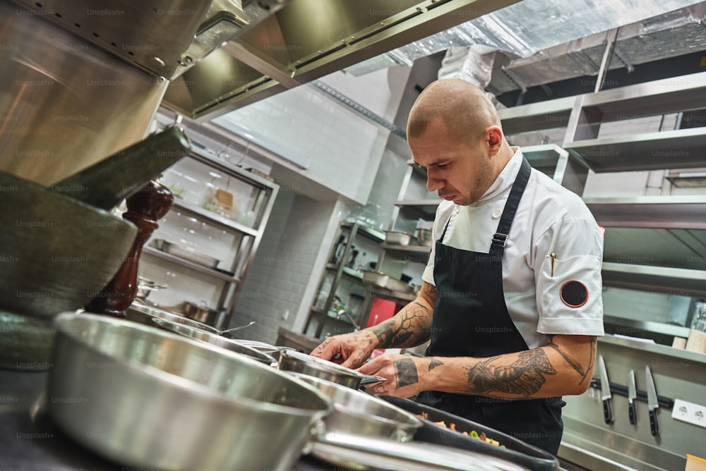Inspiration in cooking. Young male chef with several tattoos on his arms is garnishing italian pasta in a restaurant kitchen. Food decoration