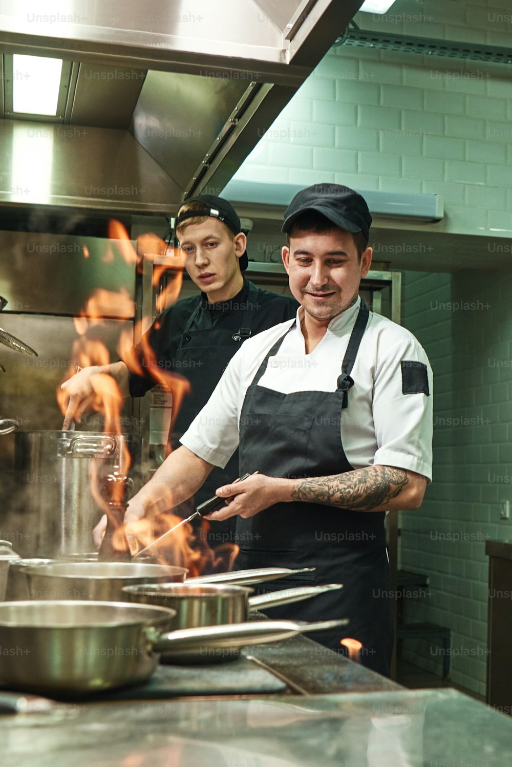 불로 일하기. 두 명의 젊고 긍정적 인 요리사가 식당 주방에서 음식에 플람베를 만드는 법을 배우고 있습니다. 요리 학교. 팀워크.