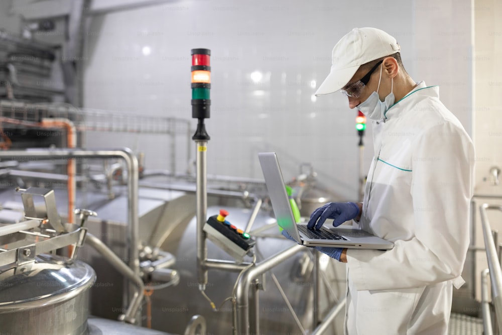 Technologist in a white coat with a laptop in his hands controls the production process in the dairy shop. Place for writing. Technologist with a laptop computer is at the factory