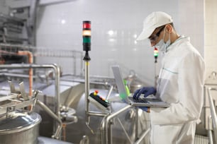 Technologist in a white coat with a laptop in his hands controls the production process in the dairy shop. Place for writing. Technologist with a laptop computer is at the factory