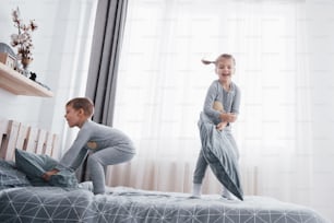 白い寝室で遊ぶ幸せな子供たち。小さな男の子と女の子、兄と妹がパジャマを着てベッドで遊んでいます。赤ちゃんと幼児のためのナイトウェアと寝具。家で家族。