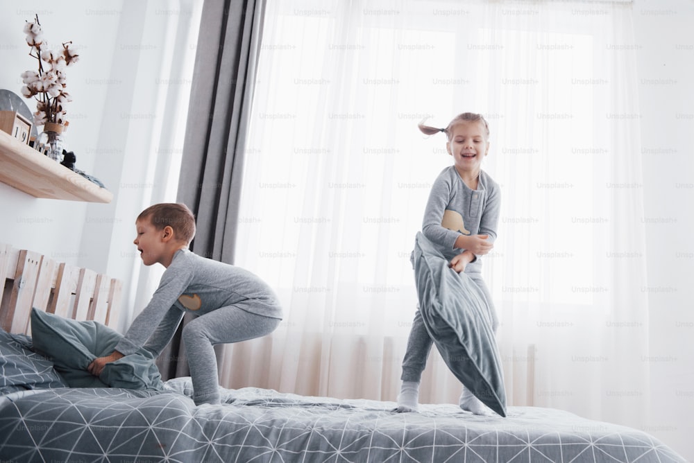 Bambini felici che giocano in camera da letto bianca. Il bambino e la bambina, il fratello e la sorella giocano sul letto indossando il pigiama. Biancheria da notte e biancheria da letto per neonati e bambini. Famiglia a casa.