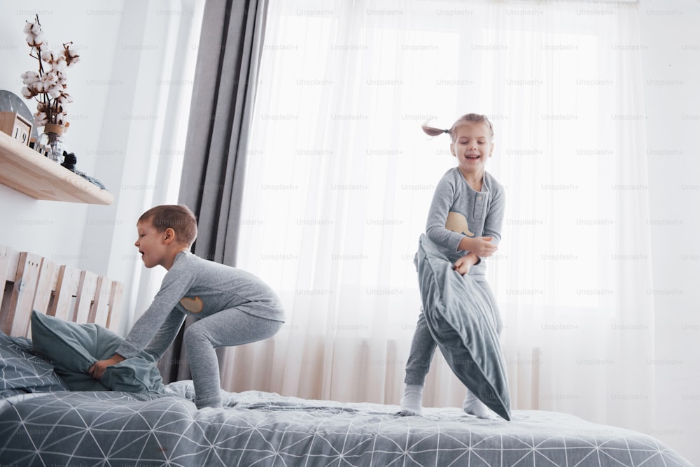 Des enfants heureux jouant dans une chambre blanche. Petit garçon et petite fille, frère et sœur jouent sur le lit en pyjama. Vêtements de nuit et literie pour bébé et tout-petit. Famille à la maison.