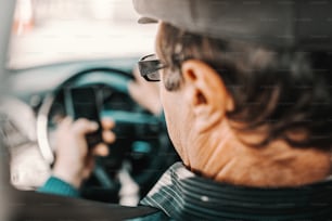Close up of serious Caucasian senior man with cap on head and eyeglasses sitting in car with hand on steering wheel and using smart phone.