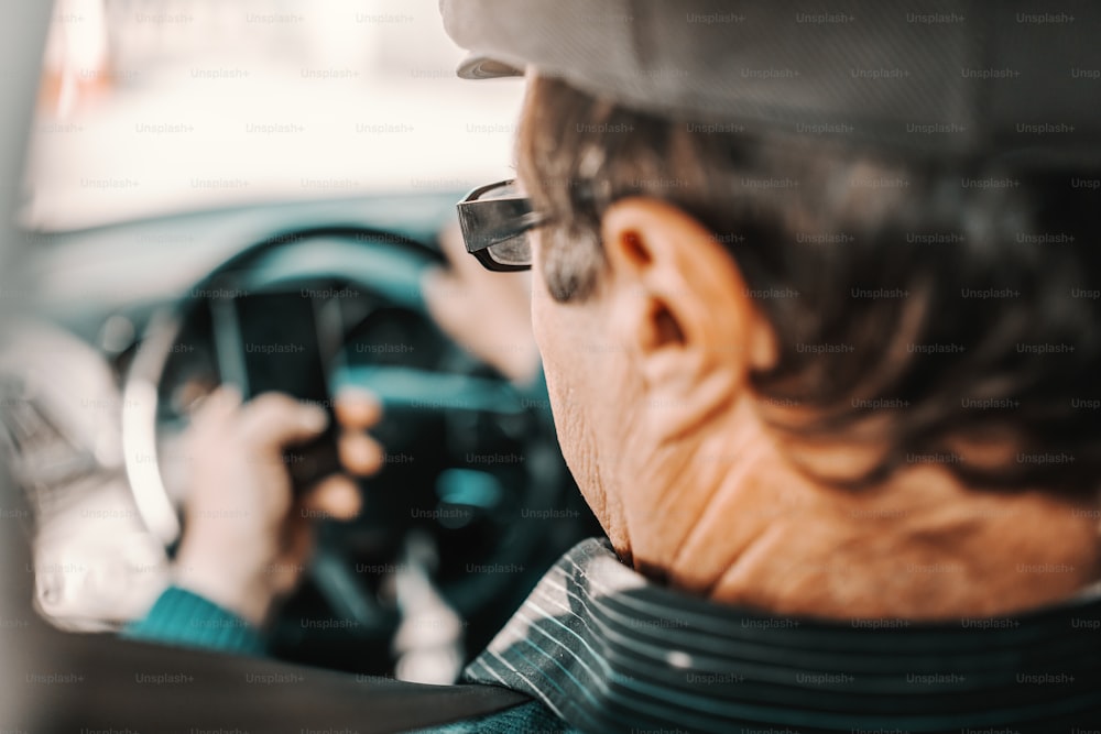 Nahaufnahme eines ernsthaften kaukasischen älteren Mannes mit Mütze auf dem Kopf und Brille, der mit der Hand am Lenkrad im Auto sitzt und ein Smartphone benutzt.