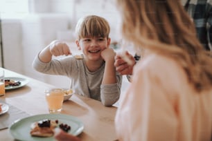 Positive kid smiling to his parents while sitting at the table with a spoonful of cornflakes in his hand