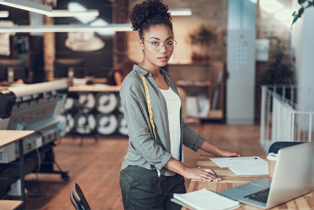 My workplace. Portrait of charming young woman with measuring tape standing near wooden table with laptop and holding pencil