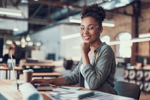 Waist up portrait of beautiful young lady sitting at wooden table with notebook and stationery. She is resting chin on hand while looking at camera and smiling