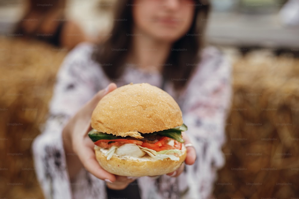Vegane Burger-Nahaufnahme in Boho-Mädchenhänden. Stylisches Hipster-Mädchen mit Sonnenbrille isst leckeren veganen Burger auf dem Street Food Festival. Glückliche Boho-Frau probiert und beißt Burger