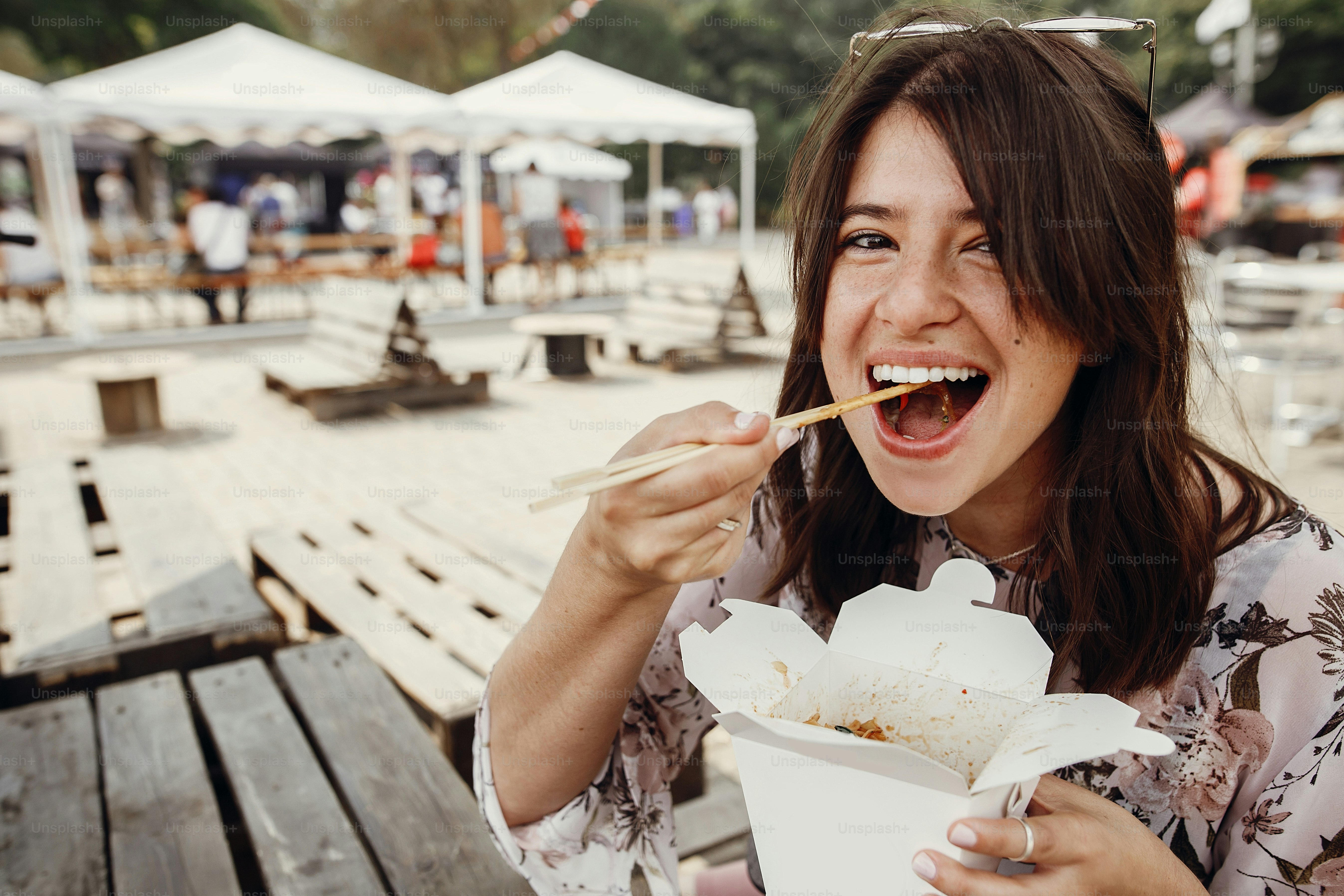 Stilvolles Hipster-Mädchen, das Wok-Nudeln mit Gemüse aus dem Karton mit Bambusstäbchen isst. Asiatisches Street Food Festival. Boho-Frau, die thailändische Nudeln in einer Papierbox zum Mitnehmen isst. Lieferung von Lebensmitteln