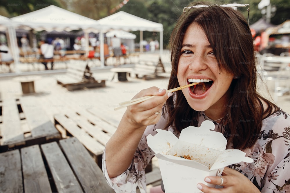 Stylish hipster girl eating wok noodles with vegetables from carton box with bamboo chopsticks. Asian Street food festival. Boho woman eating thai noodles in takeaway paper box. Food delivery